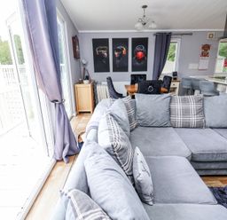A living room with a large grey sectional sofa and a dining table with chairs next to a kitchen area with bar stools and a countertop at 155 Woodland Hall Lyons in Ruthin