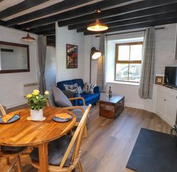 A living room with a wooden dining table and chairs croissants on plates and a vase with yellow flowers a blue sofa and an armchair a wooden chest a window with curtains a television and a fireplace at 4 Old Tanrhiw in Beddgelert