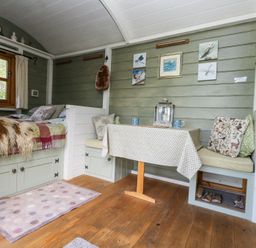 A small room with a bed, a table with a tablecloth, bench seating with cushions, wall art, and a wooden floor at Fallow in Warmwell