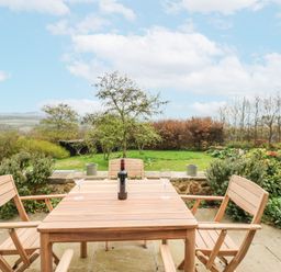 A wooden outdoor table with four chairs a bottle of wine and two glasses on a stone patio overlooking a garden at The Barn in Riding Mill near Corbridge