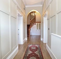 A hallway with light-colored paneled walls wooden flooring a patterned rug two doors and a staircase with a chandelier at The Burleigh in Hunstanton