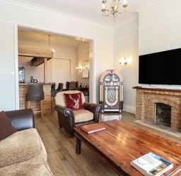 A living room with a sofa, armchair, wooden coffee table, jukebox, fireplace, and wall-mounted TV at The Burleigh in Hunstanton