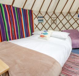 A bed with white linens and a fish pillow inside a yurt with a colorful striped blanket on the wall and a purple covered chair at Moonlight in Ventnor