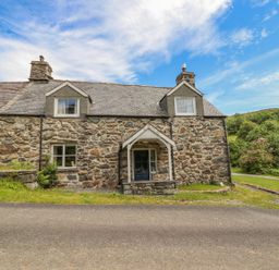 Stone cottage with two chimneys and a blue door by a paved road at Ty Glan Yr Afon in Bontddu near Dolgellau