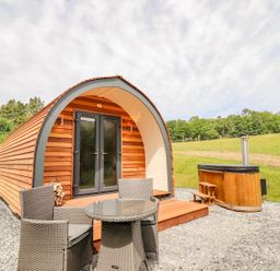 A wooden pod with double glass doors a small patio with a round table and two chairs and a hot tub outside on gravel with grass and trees behind at Briallen in Bontddu near Dolgellau