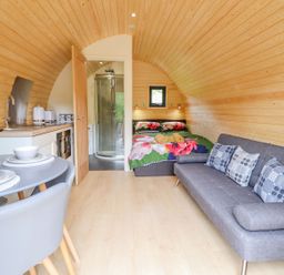 An interior view of a wooden pod with a bed floral bedding a gray sofa with cushions a small kitchen and a dining table at Briallen in Bontddu near Dolgellau