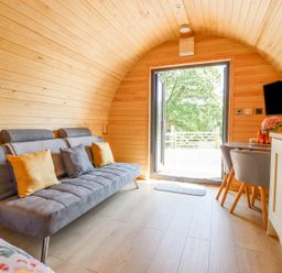 A small room with wooden curved walls a gray sofa with cushions a table with chairs a TV and an open door showing greenery outside at Eirlys in Bontddu near Dolgellau