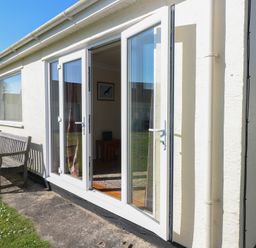 A building exterior with open glass doors next to a wooden bench and grass at Kites - 52 Gower Holiday Village in Scurlage near Port Eynon and Rhossili