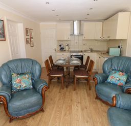 A dining area with a wooden table set for four and a kitchen with white cabinets and appliances and two blue leather armchairs with floral cushions