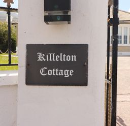 A white wall with a black rectangular sign reading Killelton Cottage next to a gate and a house in the background at Killelton Cottage in Camp County Kerry