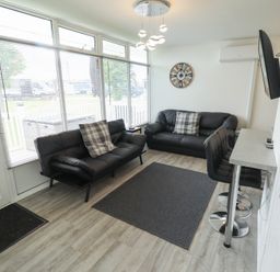A living room with two black sofas a wall clock a mounted TV and bar stools at 99 Cherry Park in Chapel St Leonards