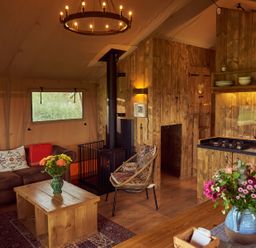 A living area with a brown leather sofa and wooden coffee table with flowers a wooden kitchen with a stove kettle and knives a woven chair and wood burning stove at Pippin lodge in Coxbridge near Glastonbury