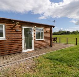 A wooden lodge with windows and a door opening onto a deck overlooking a grassy field at Twilight in Raglan