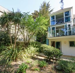 A modern house with large glass windows and balconies surrounded by various plants and trees at 6 Red Sails in Sandbanks