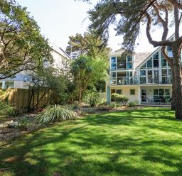 A garden with grass trees and shrubs in front of a house with large windows at 6 Red Sails in Sandbanks