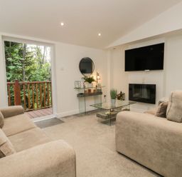 A living room with two beige sofas a glass coffee table a wall-mounted TV and a glass door opening to a wooden balcony at Mere Brook Lodge in White Cross Bay