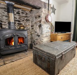 A stone fireplace with a wood-burning stove and a metal chest coffee table in front a wooden TV stand with a television and a brown leather pouf in a living room at Cobweb Cottage in Waunfawr