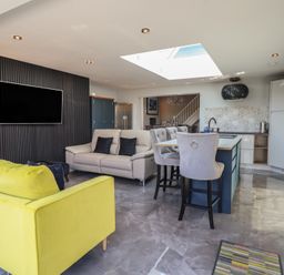 An open living room and kitchen area with yellow chairs a sofa bar stools and a wall-mounted television at Bronallt in Cemaes Bay