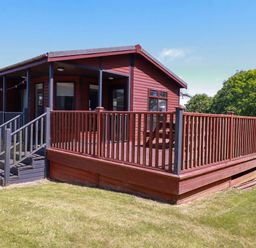 An exterior view of a wooden lodge with a fenced porch and steps leading to a grassy area at Lodge 9- Lamorna in Tregolds near St Merryn