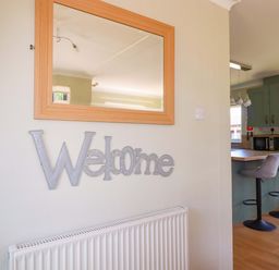 A wall with a rectangular wooden mirror and metal welcome sign above a white radiator with a kitchen area having two bar stools at Lodge 9- Lamorna in Tregolds near St Merryn