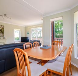 A dining table with six chairs next to a living area with black sofas a TV and windows showing trees outside at Lodge 9- Lamorna Tregolds near St Merryn