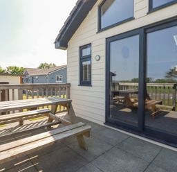 An outdoor patio with a wooden picnic table and benches next to a building with glass sliding doors and a window at Lodge 18 in St Merryn