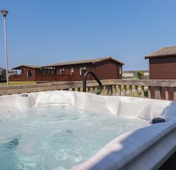 An outdoor hot tub on a deck with wooden cabins and a street lamp in the background at Lodge 18 in St Merryn