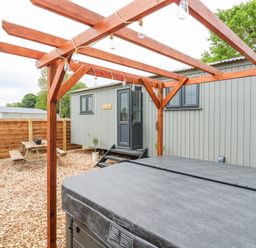 An outdoor area with a wooden pergola over a hot tub a wooden fence a picnic table and a grey wooden cabin at Woodpecker in Lode near Bottisham