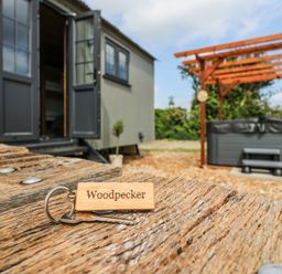A wooden key tag with the word Woodpecker on a table outside a building with open doors and a hot tub under a wooden pergola in a garden at Woodpecker in Lode near Bottisham