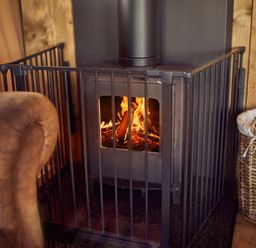 A wood stove with fire inside surrounded by a safety gate next to a leather chair and a basket of firewood at Bramley Lodge in Coxbridge near Glastonbury