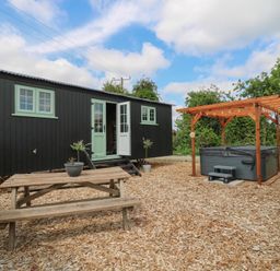 An outdoor area with a wooden picnic table potted plants a black building with green windows and doors and a hot tub under a wooden pergola at The Heron in Lode near Bottisham