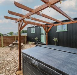 An outdoor area with a covered hot tub, a wooden pergola with hanging lights, a black cabin with a mint green door and windows, and a wooden picnic table surrounded by wood chips