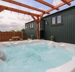 An outdoor hot tub with bubbling water under a wooden pergola next to a dark green cabin with two windows and a door at Barn Owl in Lode near Bottisham