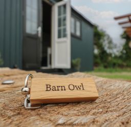 A wooden keychain with Barn Owl text on a wooden surface outside a dark green building with an open door at Barn Owl in Lode near Bottisham