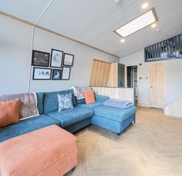 A living room with a blue sectional sofa orange ottoman wooden floor and framed pictures on the wall at Starlight Skylight - Billing Aquadrome in Billing Aquadrome Holiday Park