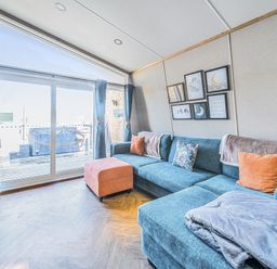 A living room with a blue sectional sofa with cushions and blankets an orange ottoman and sliding glass doors leading to a deck with a hot tub at Starlight Skylight - Billing Aquadrome Holiday Park