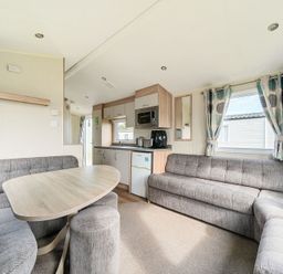 A living area with grey upholstered seating a wooden dining table a kitchen area with cabinets microwave and appliances at Golden Dune - Hayling Island in Hayling Island Holiday Park