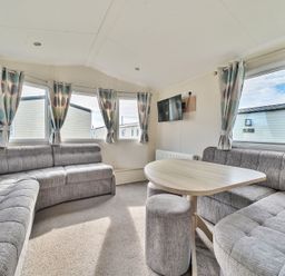A sitting area with gray sofas a wooden table a small gray stool and windows with curtains at Golden Dune - Hayling Island in Hayling Island Holiday Park