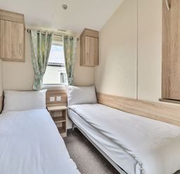 A small bedroom with two single beds separated by a wooden nightstand and a window with patterned curtains at The Lookout - Hayling Island in Hayling Island Holiday Park