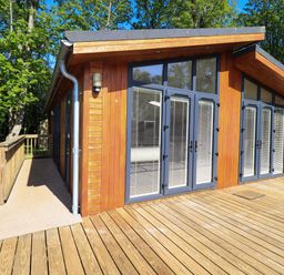 A wooden cabin with large glass doors and windows on a wooden deck surrounded by trees at Kingfisher Lodge in Gargrave