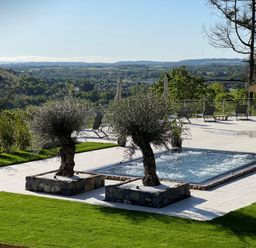 An outdoor area with two trees in stone planters next to a rectangular hot tub and several lounge chairs with umbrellas overlooking a valley at Forest View in Pontyclun