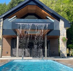 An outdoor swimming pool with water falling from a structure in front of a modern house surrounded by trees at Forest Lodge in Pontyclun