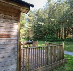 An outdoor wooden deck with railing and a barbecue grill next to a wooden building with a sycamore sign surrounded by trees at Sycamore Lodge in Rosliston