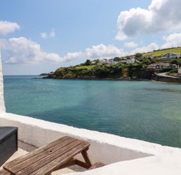 A balcony with a wooden table and storage box overlooking a coastal view with houses on a hill at Rock Cottage Portmellon near Mevagissey