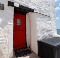 A red door set in a white stone wall next to a black outdoor storage box by the sea at Rock Cottage Portmellon near Mevagissey