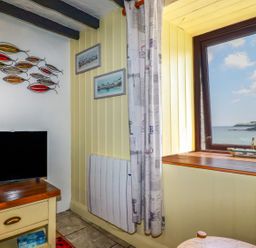A corner of a room with a television on a wooden table metal fish wall art two framed pictures a window overlooking the sea and a curtain at Rock Cottage Portmellon near Mevagissey