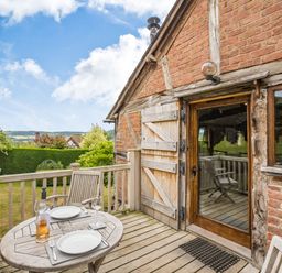 An outdoor wooden deck with a table set for two and wooden chairs next to a brick and timber house at The Cider Mill in Westhope near Leominster
