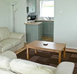 A living room with two white sofas a wooden coffee table a floor lamp and a kitchen area in the background at Seascape in Kessingland near Lowestoft
