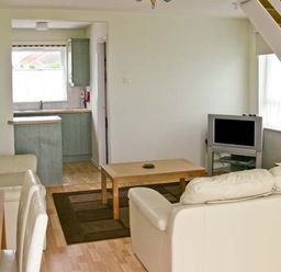 A living room with two cream sofas a wooden coffee table a TV on a stand and a kitchen with green cabinets visible at Seascape in Kessingland near Lowestoft