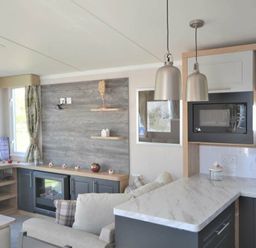 a living room with a grey sofa, a TV unit, a kitchenette with marble counter, and decorative shelves at Perran Sands - Holiday Accommodation 15554 in Perranporth